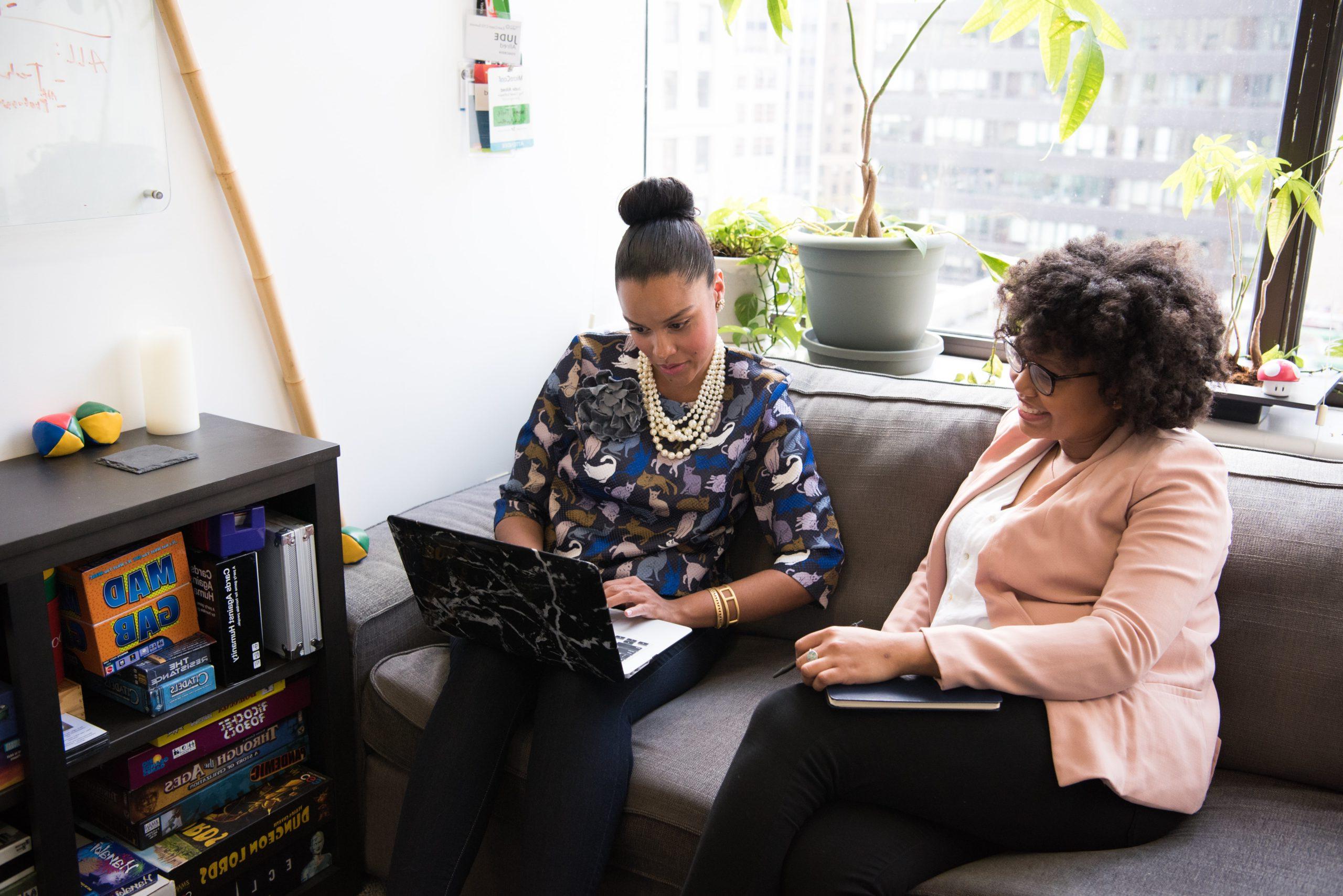 Two women sitting on a couch together looking at a laptop