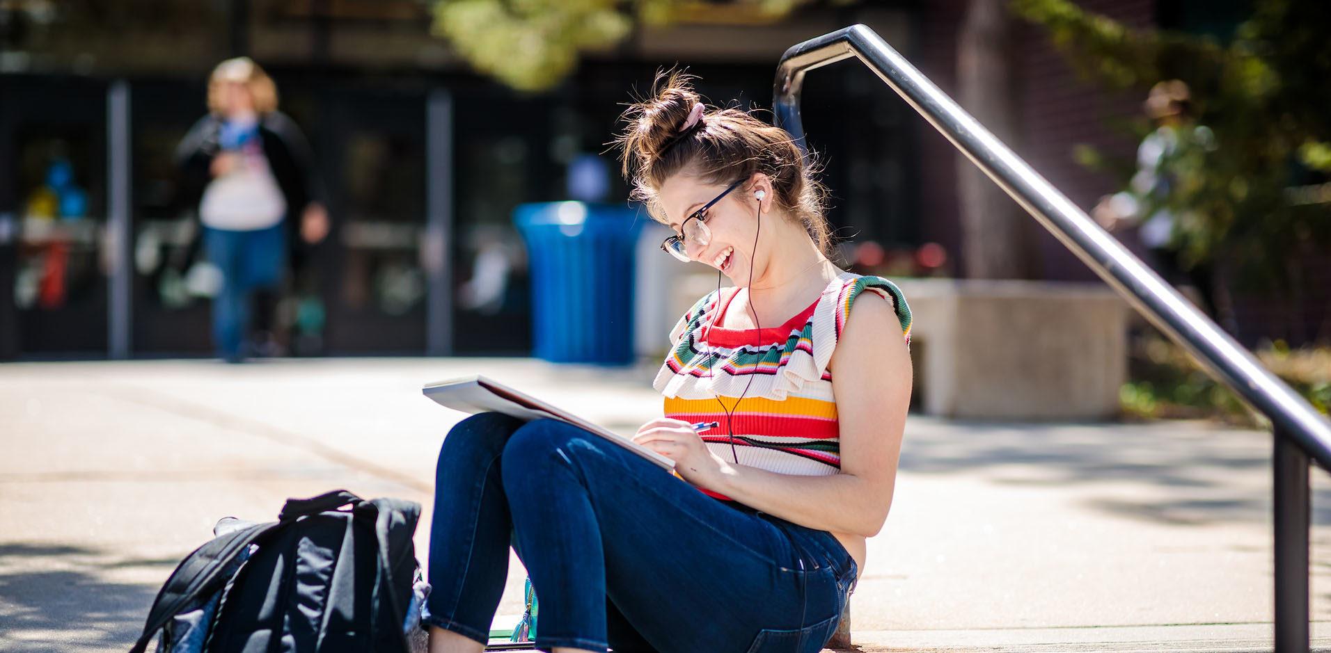 Student sitting on outside stairs writing in notebook and wearing headphones