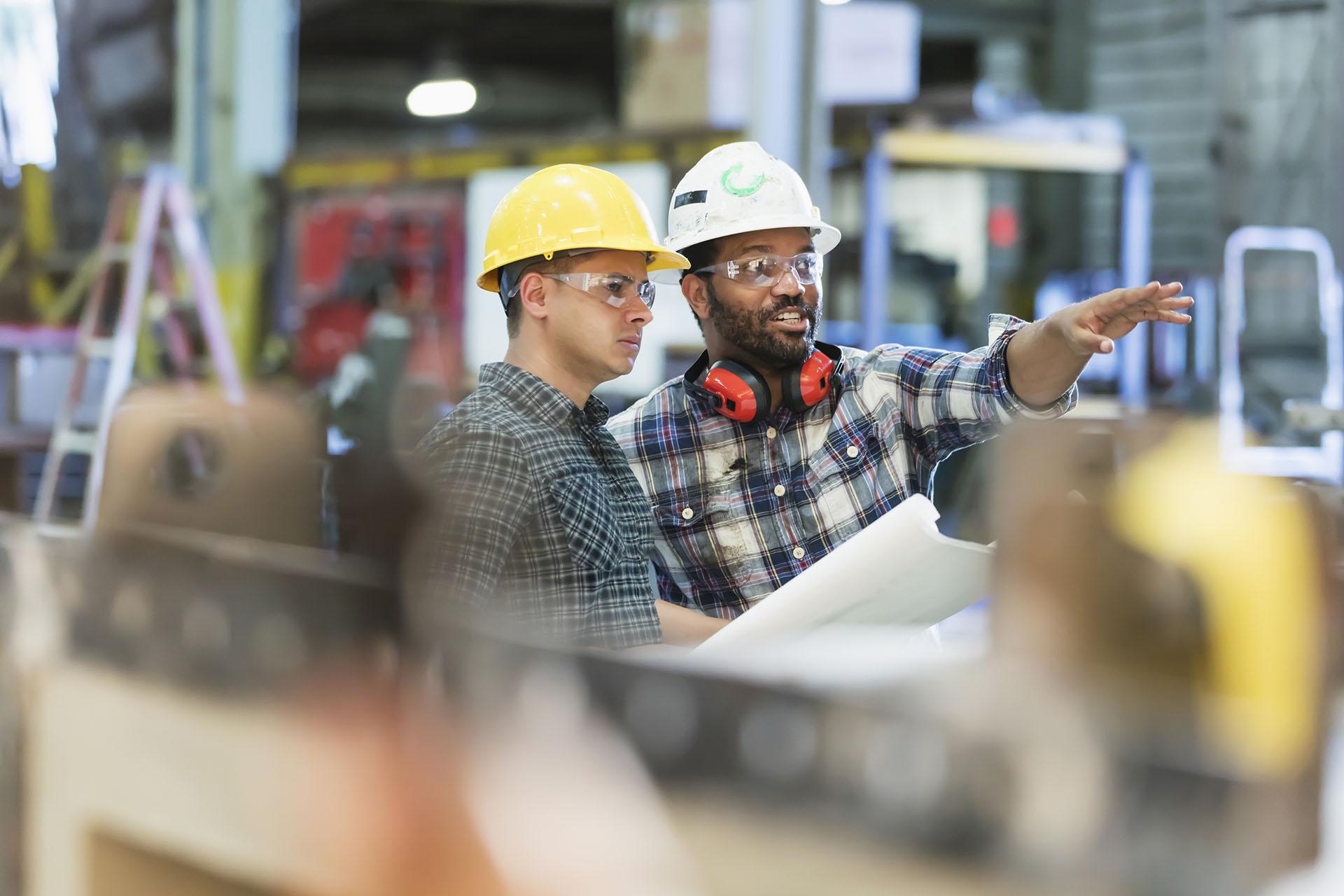 Two multi-ethnic workers in their 30s talking in a metal fabrication plant wearing hardhats and protective eyewear. The man pointing is African-American and his coworker is Hispanic.